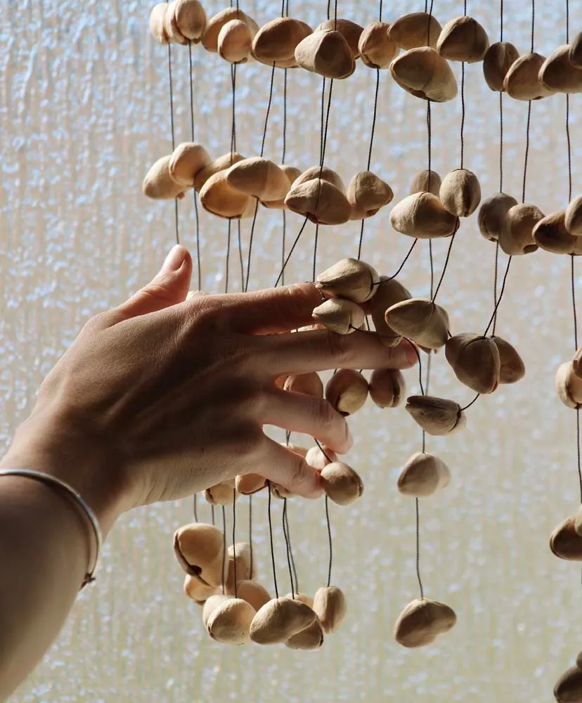 A hand gently reaching toward a hanging chime of natural seed pods by a bright window