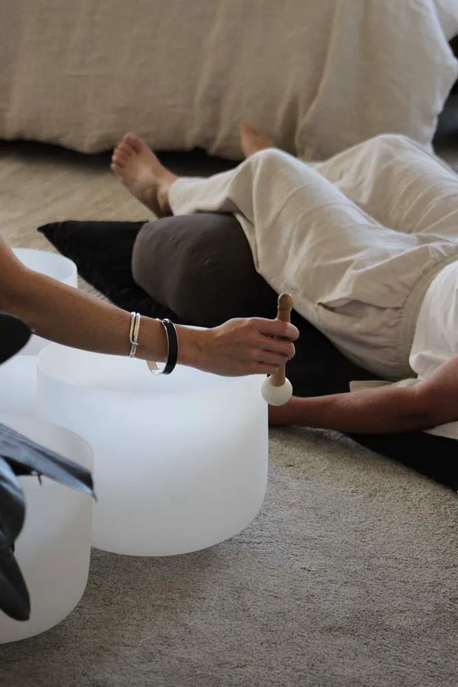 Hands playing a crystal singing bowl beside a reclining participant